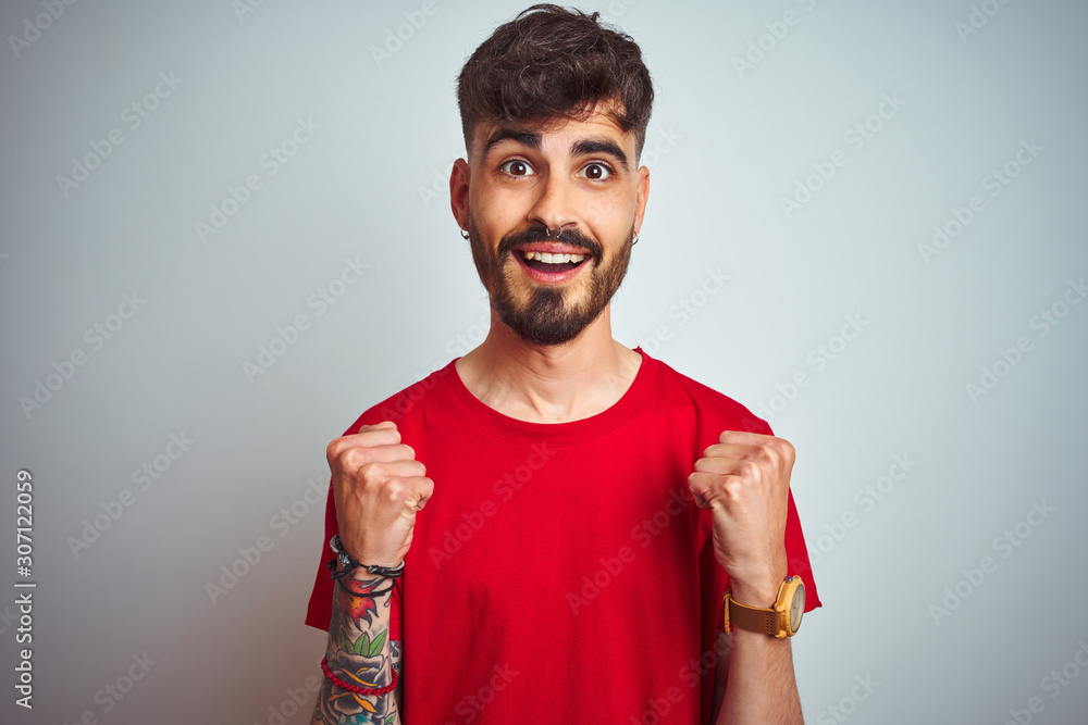 Young man with tattoo wearing red t-shirt standing over isolated white background celebrating surprised and amazed for success with arms raised and open eyes. Winner concept. Young man with tattoo wearing red t-shirt standing over isolated white background celebrating surprised and amazed for success with arms raised and open eyes. Winner concept.