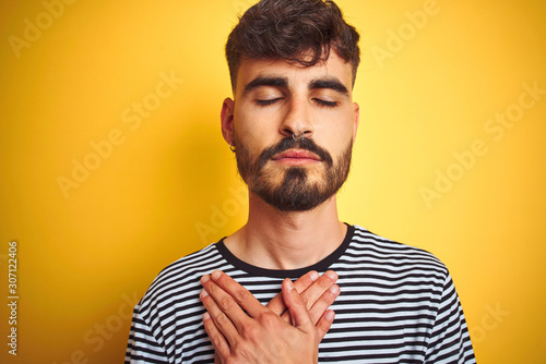 Young man with tattoo wearing striped t-shirt standing over isolated yellow background smiling with hands on chest with closed eyes and grateful gesture on face. Health concept. © Krakenimages.com