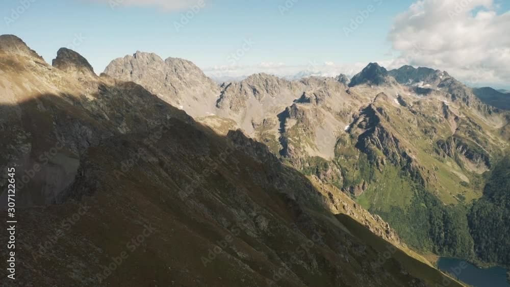 Aerial view; drone flying over mountain range Loub, Kardyvach lake at the foot; rocky peaks of mounts, sunlight shining through the clouds; steep stony slopes, dangerous hiking route, Krasnaya Polyana Aerial view; drone flying over mountain range Loub, Kardyvach lake at the foot; rocky peaks of mounts, sunlight shining through the clouds; steep stony slopes, dangerous hiking route, Krasnaya Polyana