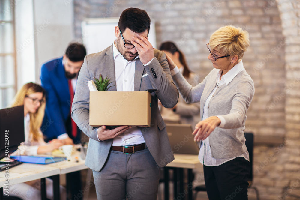 Handsome businessman in light modern office with carton box. Junior office worker gets fired by senior female manager. Last day at work. Handsome businessman in light modern office with carton box. Junior office worker gets fired by senior female manager. Last day at work.