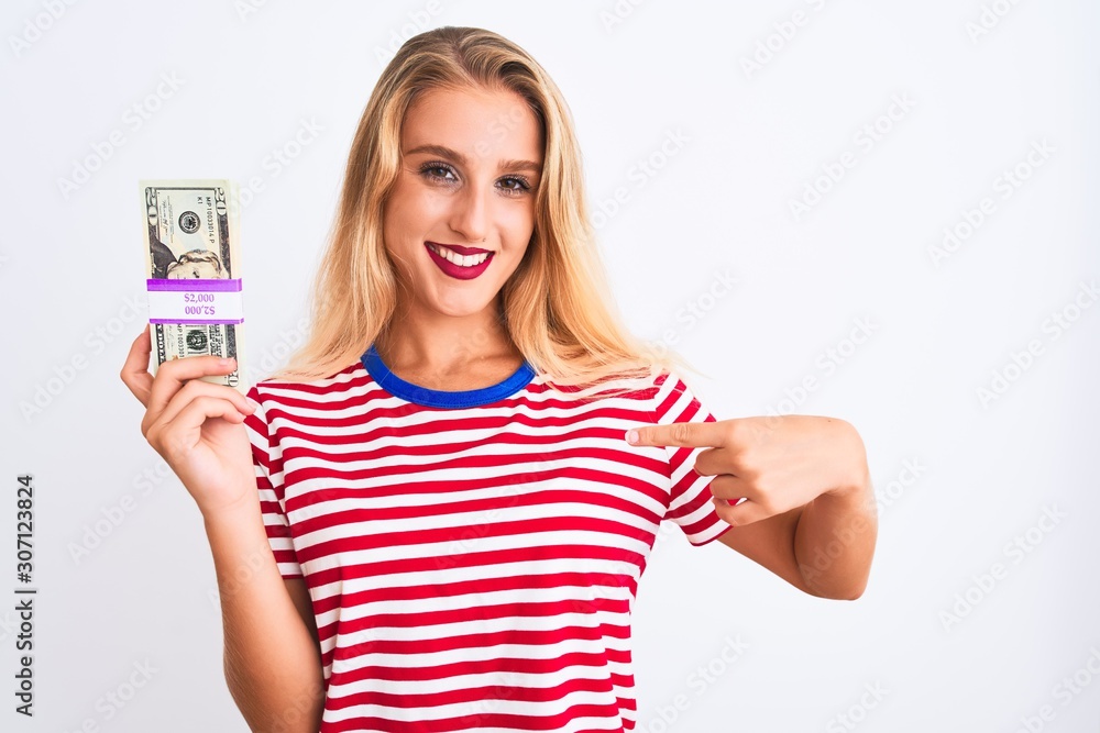 Beautiful woman wearing red striped t-shirt holding dollars over isolated white background with surprise face pointing finger to himself Beautiful woman wearing red striped t-shirt holding dollars over isolated white background with surprise face pointing finger to himself