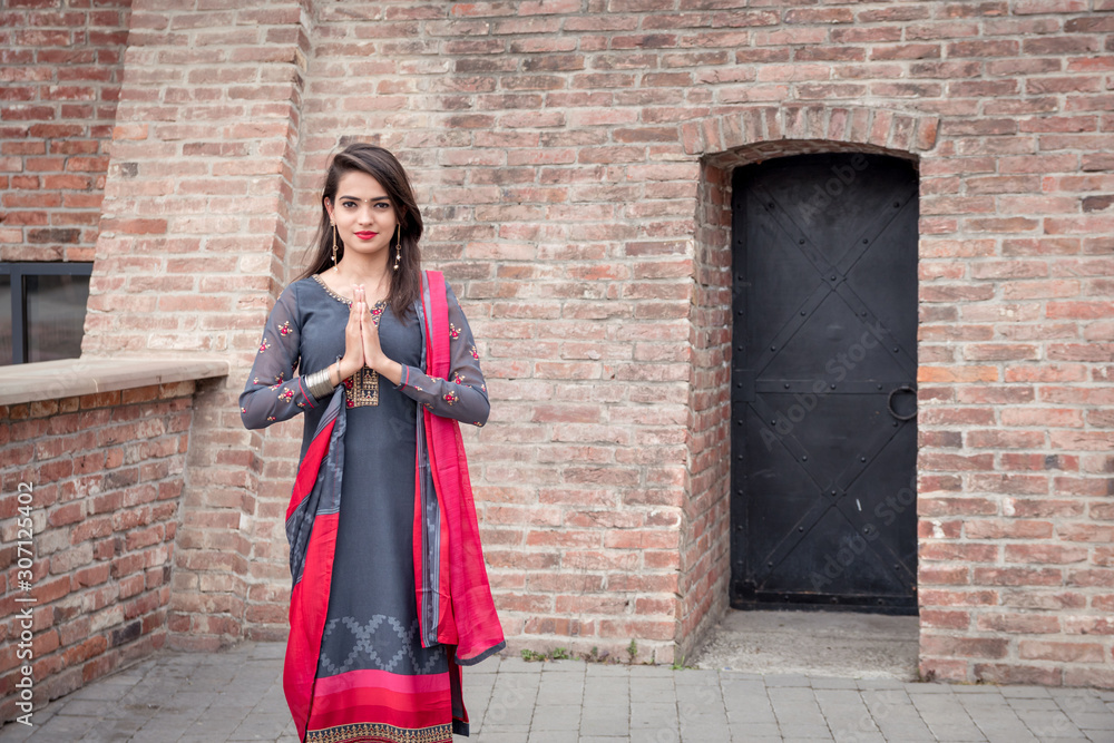 Smiling ethnic girl, hand in traditional indian mudra gesture stand. Girl in traditional Indian clothing, salwar kameez.  Indian girl in traditional dress stand at the entrance to the house.