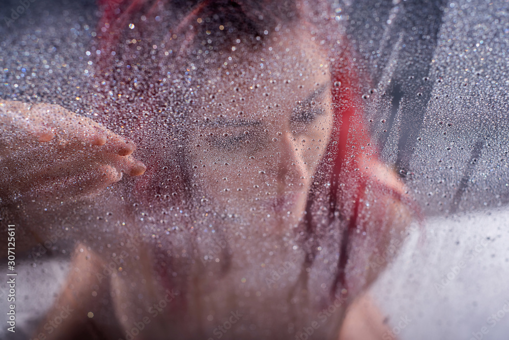 The red-haired woman takes a shower. A girl covering her breasts with her hands stands behind a misted glass.