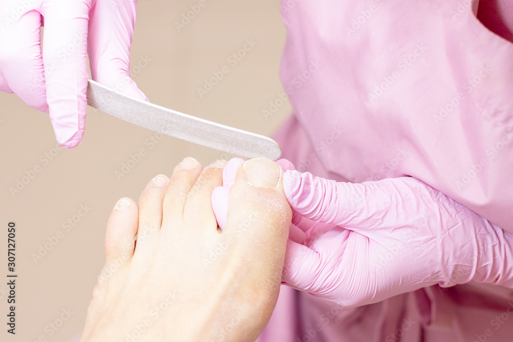 Young woman getting professional pedicure in a beauty salon, closeup. Hands a pedicurist in protective rubber gloves are applied with nails to the nails using a nail file. Professional beauty salon. Young woman getting professional pedicure in a beauty salon, closeup. Hands a pedicurist in protective rubber gloves are applied with nails to the nails using a nail file. Professional beauty salon.