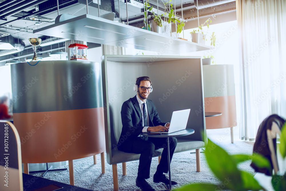 Smiling handsome caucasian bearded businessman in suit, with eyeglasses and headphones sitting at workplace and typing on laptop while looking at camera. Smiling handsome caucasian bearded businessman in suit, with eyeglasses and headphones sitting at workplace and typing on laptop while looking at camera.