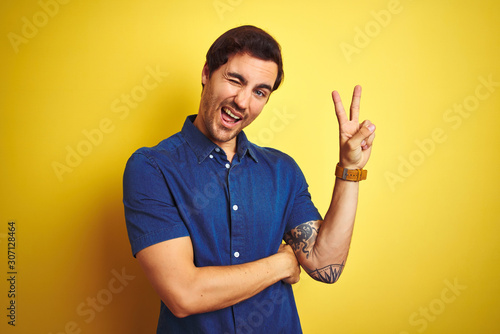 Young handsome man with tattoo wearing casual shirt standing over isolated yellow background smiling with happy face winking at the camera doing victory sign. Number two.