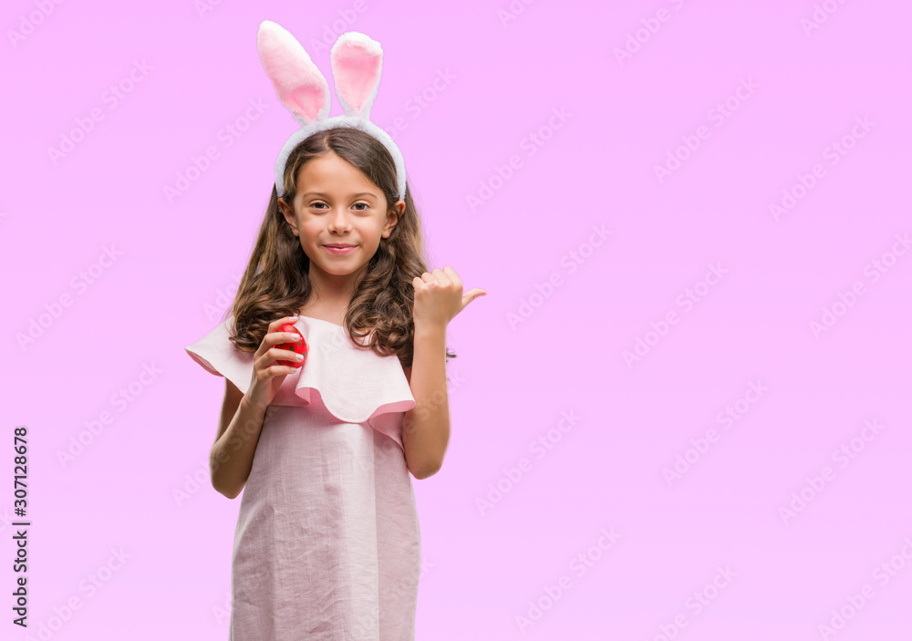 Brunette hispanic girl wearing easter rabbit ears pointing and showing with thumb up to the side with happy face smiling Brunette hispanic girl wearing easter rabbit ears pointing and showing with thumb up to the side with happy face smiling