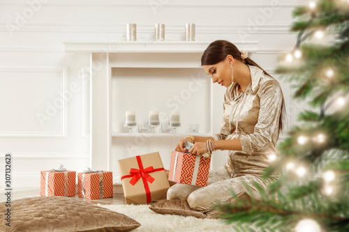 Brunette sits on carpet in front of ecological fireplace.The interior of white apartment and Christmas tree.Dressed in comfortable clothes,she unpacks gifts.Morning light coming through the window.