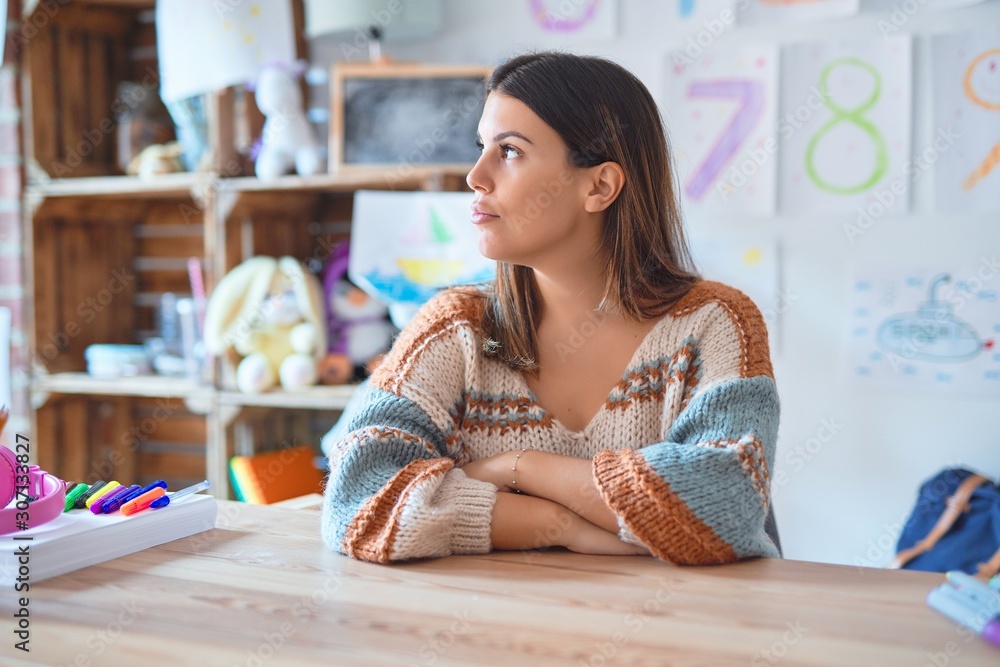 Young beautiful teacher woman wearing sweater and glasses sitting on desk at kindergarten looking to side, relax profile pose with natural face with confident smile. Young beautiful teacher woman wearing sweater and glasses sitting on desk at kindergarten looking to side, relax profile pose with natural face with confident smile.