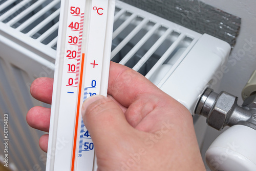 Thermometer in the hand of a person measuring the temperature near a heating radiator. Celsius temperature scale. Winter heating, concept image.