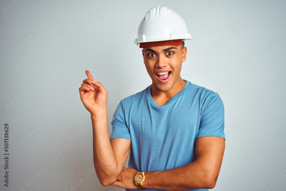 Young brazilian engineer man wearing security helmet standing over isolated white background with a big smile on face, pointing with hand and finger to the side looking at the camera.