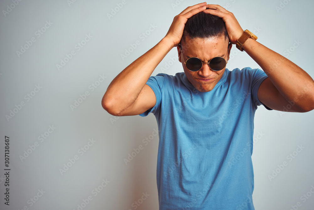 Young brazilian man wearing blue t-shirt and sunglasses over isolated white background suffering from headache desperate and stressed because pain and migraine. Hands on head. Young brazilian man wearing blue t-shirt and sunglasses over isolated white background suffering from headache desperate and stressed because pain and migraine. Hands on head.