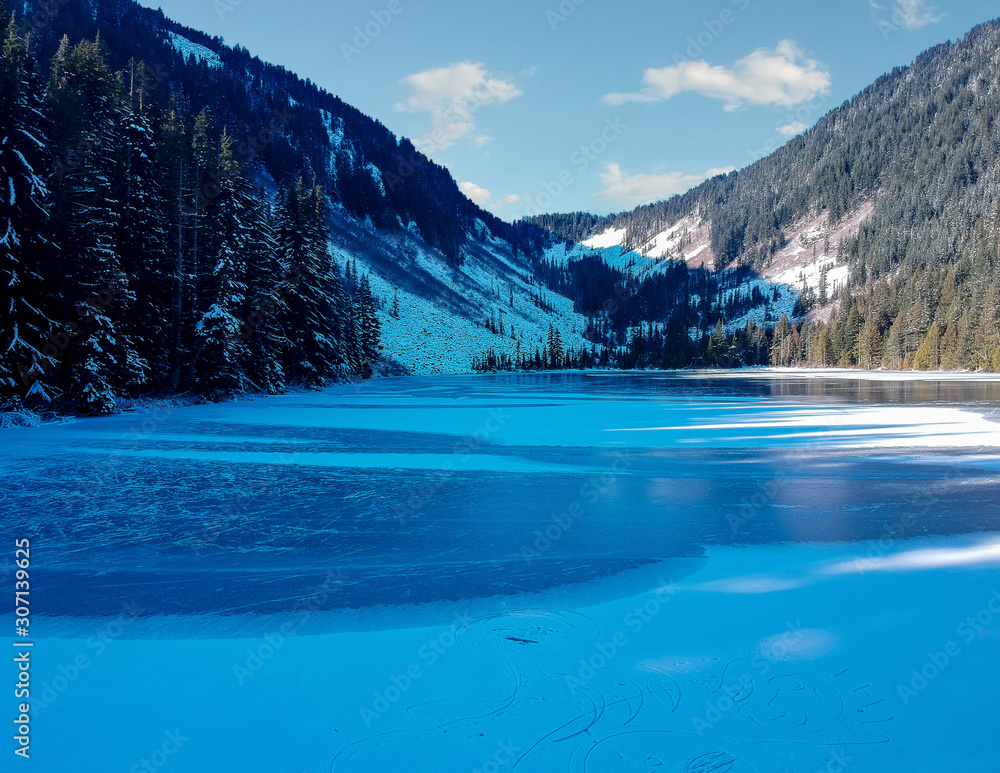 Frozen partially shaded Talapus Lake of the Alpine Lake Wilderness on a snow covered day with Cedars and Hemlock trees on the mountain side in Washington State.