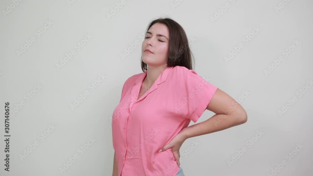 Sick young woman isolated on gray background in studio having pain in back, looking dissatisfaction in casual pink shirt. People sincere emotions, lifestyle concept. Sick young woman isolated on gray background in studio having pain in back, looking dissatisfaction in casual pink shirt. People sincere emotions, lifestyle concept.