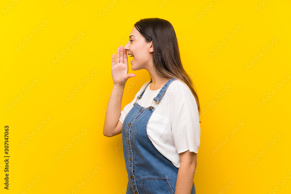 Young woman in dungarees over isolated yellow background shouting with mouth wide open to the lateral Young woman in dungarees over isolated yellow background shouting with mouth wide open to the lateral