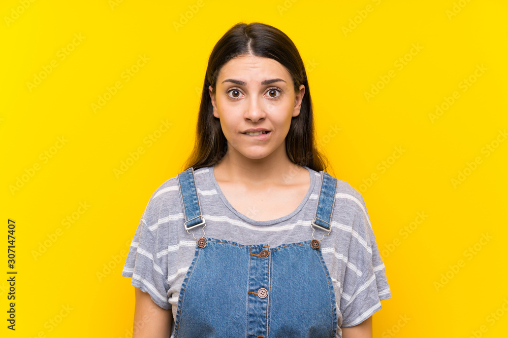 Young woman in dungarees over isolated yellow background having doubts and with confuse face expression Young woman in dungarees over isolated yellow background having doubts and with confuse face expression