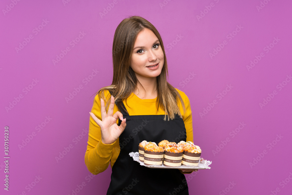 Teenager girl holding lots of different mini cakes over isolated purple background showing an ok sign with fingers Teenager girl holding lots of different mini cakes over isolated purple background showing an ok sign with fingers