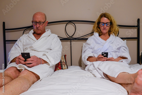 Tbilisi, Georgia A middle aged couple in bed with white bathrobes reading their phones.
