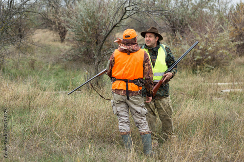 A mans with a gun in his hands and an orange vest on a pheasant hunt in a wooded area in cloudy weather. Hunters with dogs in search of game.