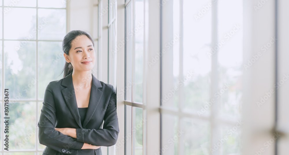 Fototapeta premium Portrait of smiling pretty young business woman crossed her arms and standing on workplace office and looking out to window.Smile and Thinking for future.