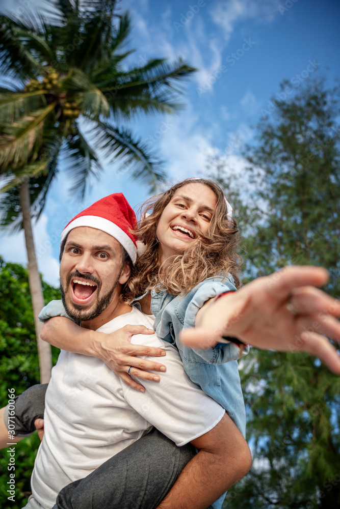 Fototapeta premium Young beautiful couple in Santa Claus hats walks along the shore of the tropical sea at the resort, vacation and travel for the New Year and Christmas time