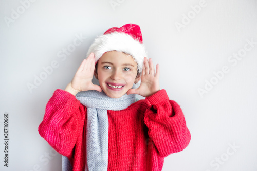 7 years old child in santa hat  red pullover and gray scarf smiles with hands near head - opened eyes to see christmas and new year presents. One  isolated  space for text