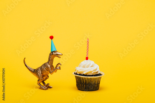 Fototapeta Naklejka Na Ścianę i Meble -  Toy dinosaur in party cap and cupcake with candle on yellow background