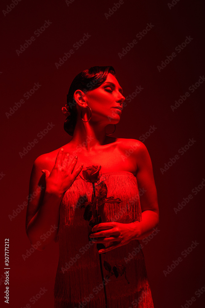 elegant tango dancer with closed eyes holding red rose on dark background with red lighting