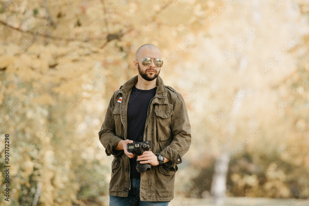 Bald photographer with a beard in aviator sunglasses with mirror lenses, olive cargo combat military jacket, blue shirt with digital wristwatch holds his camera in the forest in the afternoon.