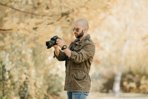 Bald photographer with a beard in aviator sunglasses with mirror lenses, olive cargo military jacket, blue jeans and shirt with digital wristwatch looking for the photo in the camera in the forest