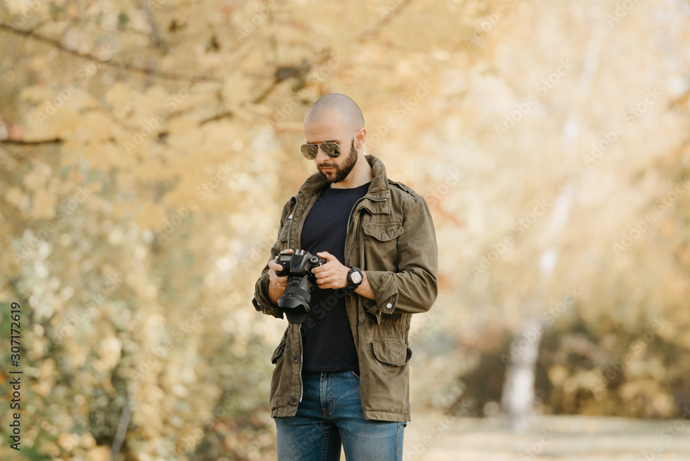 Bald photographer with a beard in aviator sunglasses with mirror lenses, olive cargo military jacket, blue jeans and shirt with digital wristwatch checks photos in the camera in the forest. Bald photographer with a beard in aviator sunglasses with mirror lenses, olive cargo military jacket, blue jeans and shirt with digital wristwatch checks photos in the camera in the forest.