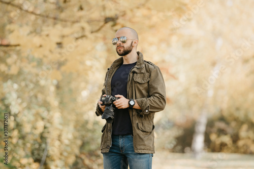 Bald photographer with a beard in aviator sunglasses with mirror lenses, olive military combat jacket, blue jeans and shirt with digital wristwatch holds the camera and looks straight in the forest.