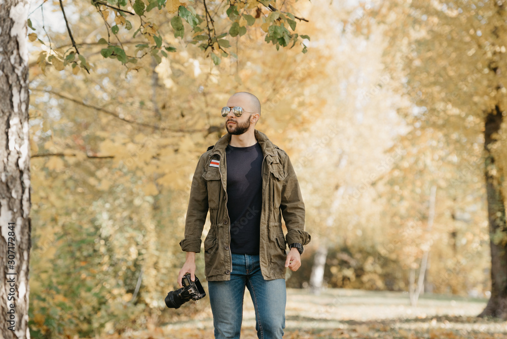 Naklejka premium Bald photographer with a beard in aviator sunglasses with mirror lenses, olive military combat jacket, blue jeans and shirt with digital wristwatch holds the camera and goes straight in the park