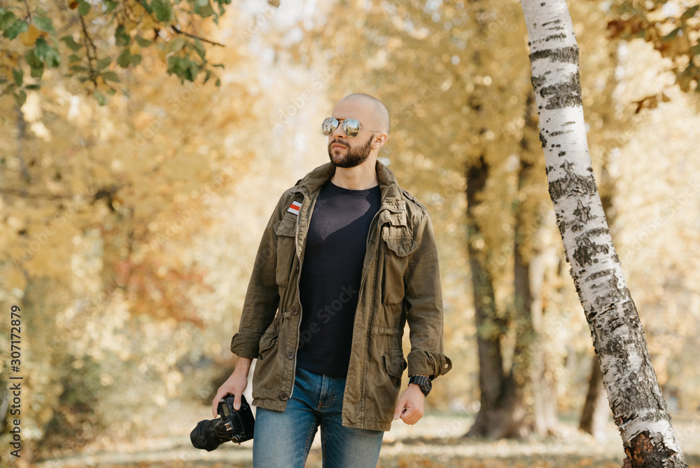 Bald photographer with a beard in aviator sunglasses with mirror lenses,  olive military jacket, jeans and shirt with digital wristwatch holds the  DSLR camera walks near the battlefield in the forest. Stock