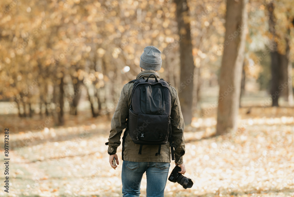 A journalist with a beard in olive military combat jacket, jeans, hat with backpack and wristwatch holds the DSLR camera and the smartphone goes straight. Portrait from the back.