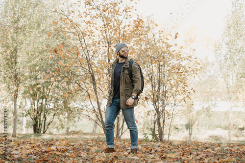 A brave traveler with a beard in aviator sunglasses with mirror lenses, olive military combat jacket, jeans, hat with backpack and wristwatch walks in the afternoon in the forest. A photojournalist.  © Roman Tyukin