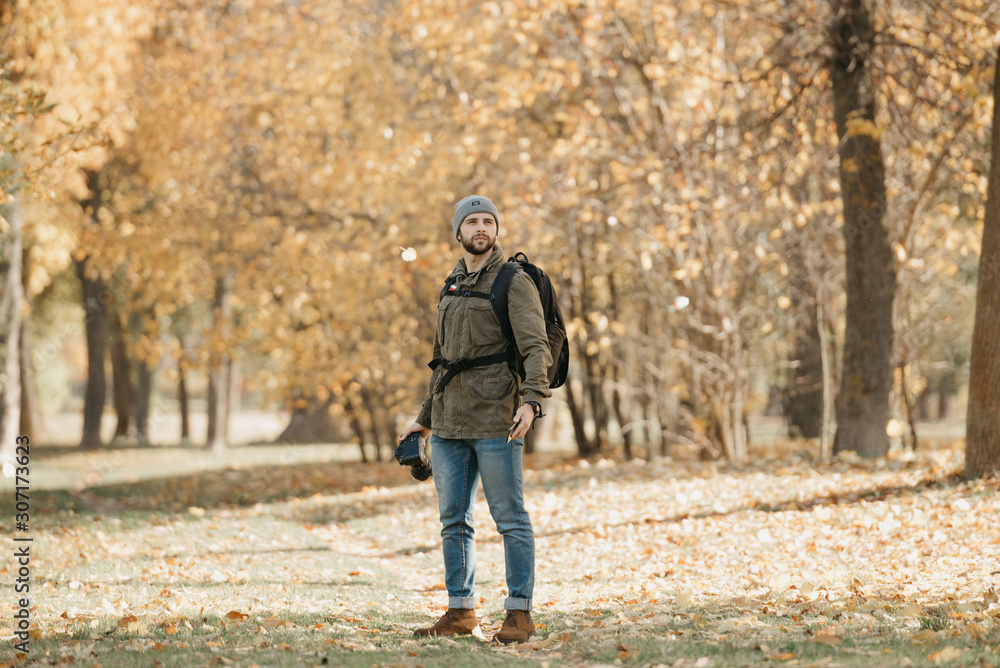 Fototapeta premium A photographer with a beard in olive military cargo combat jacket, jeans, hat with backpack and wristwatch holds the DSLR camera and the smartphone in the forest at the noon.