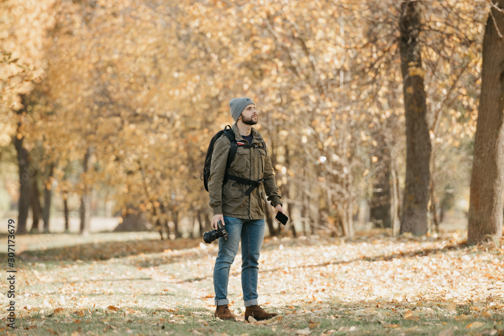 A photographer with a beard in olive military cargo combat jacket, jeans, hat with backpack and wristwatch holds the DSLR camera, the smartphone and waits in the forest at the noon. A photographer with a beard in olive military cargo combat jacket, jeans, hat with backpack and wristwatch holds the DSLR camera, the smartphone and waits in the forest at the noon.