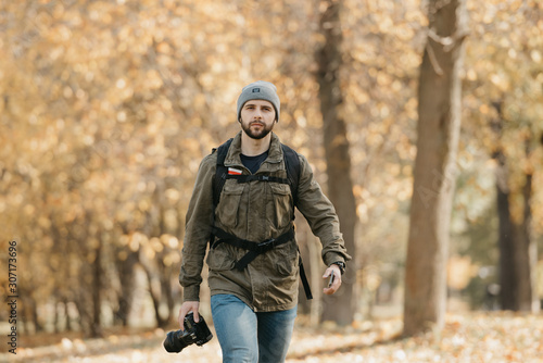 A photographer with a beard in olive military combat cargo jacket, jeans, hat with backpack and wristwatch holds the DSLR camera and the smartphone goes straight at the afternoon. © Roman Tyukin
