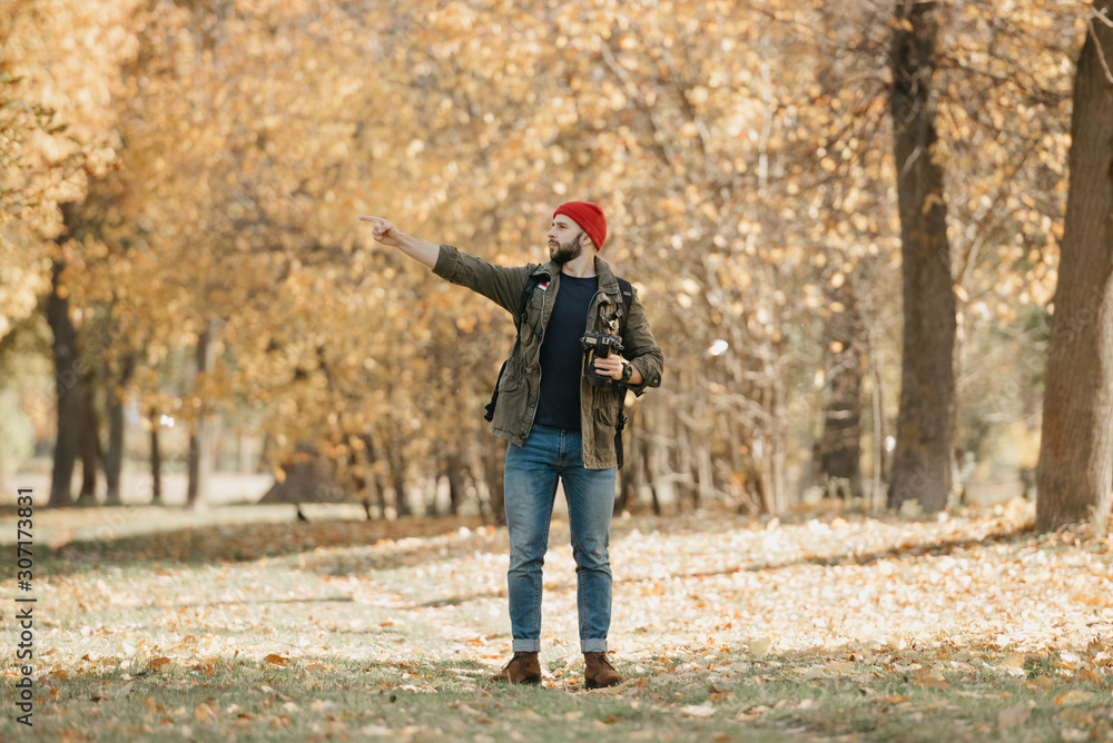 A photographer with a beard in olive military combat jacket, jeans, red hat with backpack and wristwatch holds the professional DSLR camera points a finger to the left in the forest at the afternoon  