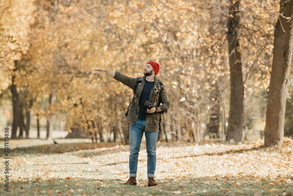 A photographer with a beard in olive military combat jacket, jeans, red hat with backpack and wristwatch holds the DSLR camera points a hand to the left in the forest at the afternoon