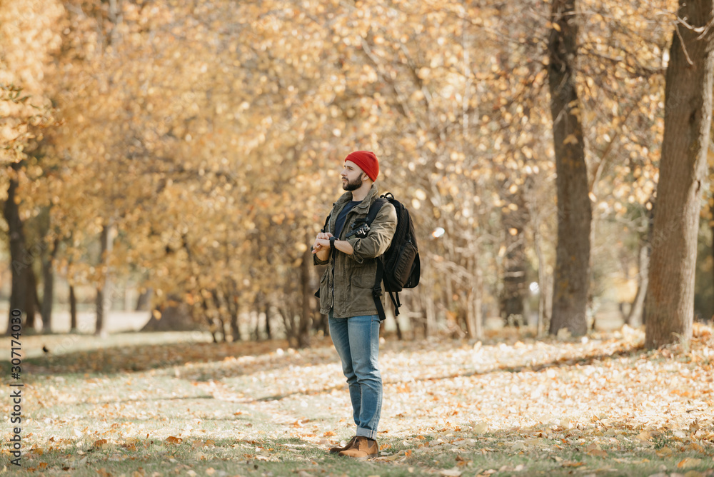 A photographer with a beard in olive military combat cargo jacket, jeans, red hat with backpack holds the DSLR camera and his wristwatch and looks away in the forest at the afternoon A photographer with a beard in olive military combat cargo jacket, jeans, red hat with backpack holds the DSLR camera and his wristwatch and looks away in the forest at the afternoon