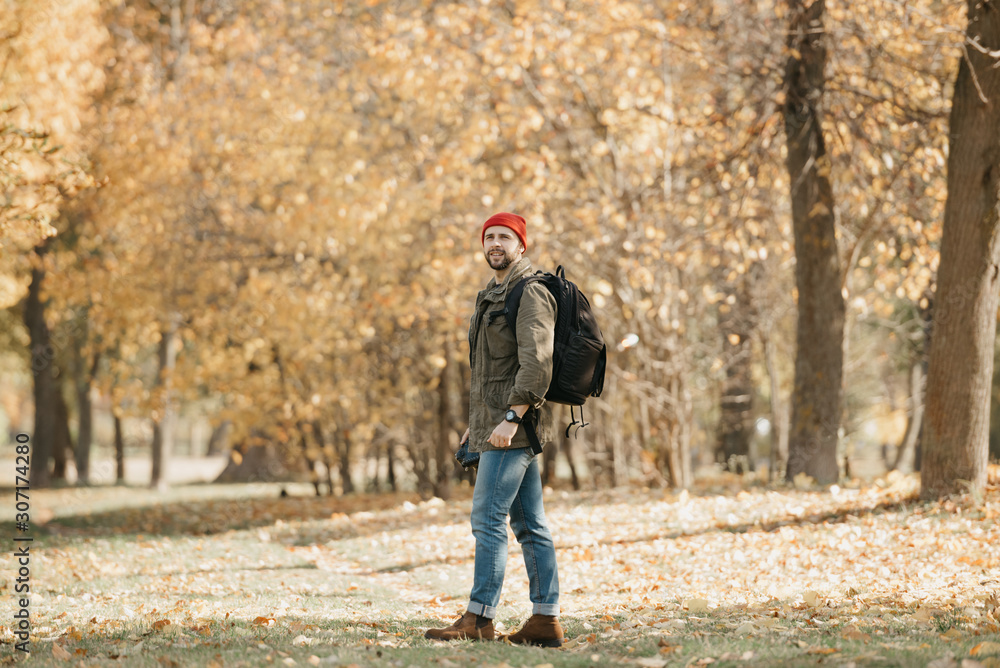 A photojournalist with a beard in an olive military cargo jacket, jeans, red hat with backpack and wristwatch holds his professional DSLR camera and waits in the forest at the noon A photojournalist with a beard in an olive military cargo jacket, jeans, red hat with backpack and wristwatch holds his professional DSLR camera and waits in the forest at the noon