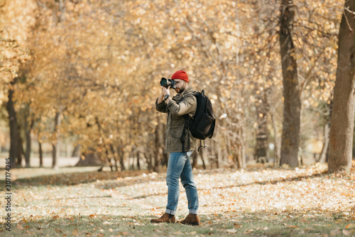 A photojournalist with a beard in an olive military cargo jacket, jeans, red hat with backpack and wristwatch takes photos with his professional DSLR camera in the forest at the noon © Roman Tyukin