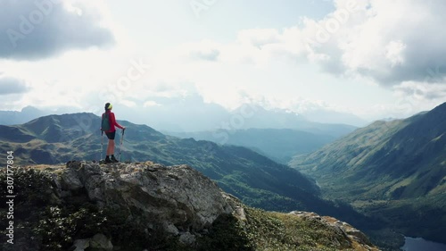 Aerial view; drone flies near tourist with hiking poles standing on summit; beautiful mountain landscape with green forest and river valleys on background; silhouette of athletic woman on sunset