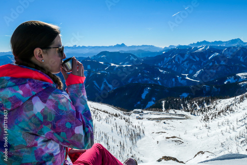 A snowboarding girl sitting at the mountain edge in Goldeck, Austria and sipping tea from a thermal bottle. Girl is enjoying the panoramic view on snow caped Alps. Happiness and serenity photo