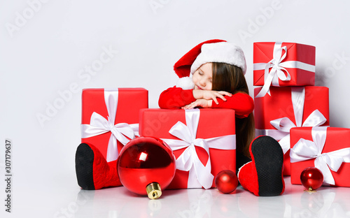 Cute little kid girl in red sweater, red cap felt boots is sitting dreaming surrounded by Christmas gifts red boxes