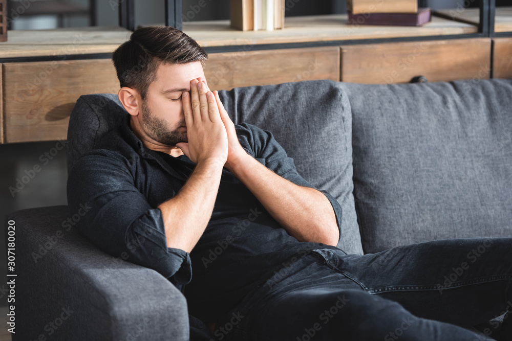 man with headache sitting on sofa and obscuring face in apartment man with headache sitting on sofa and obscuring face in apartment