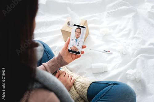 cropped view of ill woman with pills and thermometer having online consultation with male doctor on smartphone
