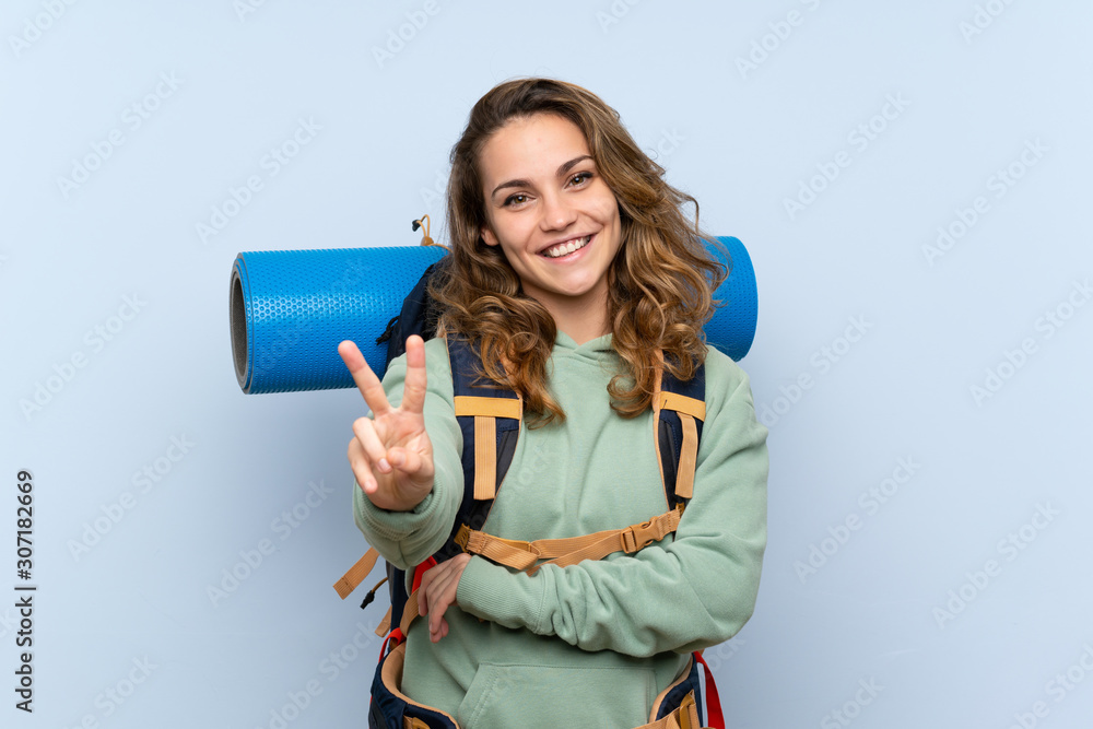 Naklejka premium Young blonde hiker girl over isolated blue background smiling and showing victory sign